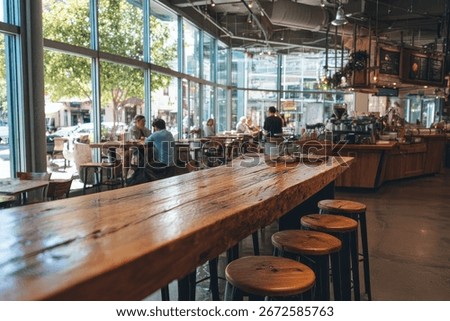 A long, rustic wooden table dominates the foreground of a bright, modern cafe. Patrons are seated at smaller tables near large windows overlooking a city str...