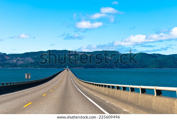 Long scenic road bridge with several lanes with\
raised sections and fencing in wide mouth of the Columbia River in\
Astoria, Pacific, rests on the horizon on the opposite bank of the\
hilly with trees.