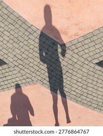 Long shadow of person on patterned pavement. Pink and gray tiles form geometric design. Shadow stretches across textured ground surface. Person stands at intersection of distinct paving sections