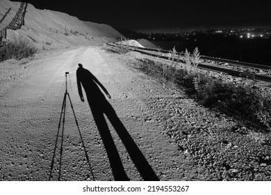 Long Shadows Of A Photographer And Camera Mounted On A Tripod, From An Industrial Spotlight On A Rocky Road In A Mining Facility, With City Lights In The Background. A Nighttime Black And White Image.