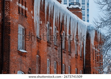 Long, sharp icicles are hanging from the snow-covered roof of an old brick building, creating a beautiful yet dangerous winter scene with a modern building in the background