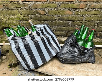 A long shot capturing several discarded green and brown glass bottles collected for recycling purposes. The empty beverage bottles are packed tightly inside two different plastic carriers: one black-and-white vertically striped plastic bag and one plain black trash bag. They are placed outdoors on a weathered wooden board against a rustic, old red brick wall covered in green moss and rough cement mortar.