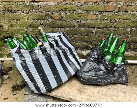 A long shot capturing several discarded green and brown glass bottles collected for recycling purposes. The empty beverage bottles are packed tightly inside two different plastic carriers: one black-and-white vertically striped plastic bag and one plain black trash bag. They are placed outdoors on a weathered wooden board against a rustic, old red brick wall covered in green moss and rough cement mortar.