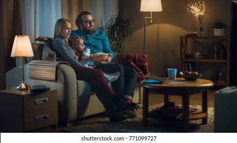 Long Shot of a Father, Mother and Little Girl Watching TV. They Sit on a Sofa in Their Cozy Living Room and Eat Popcorn. It's Evening.