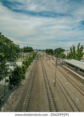A long shot of a railway track stretching into the horizon under a blue sky with white clouds. The tracks are flanked by green trees and a small train station can be seen in the distance.