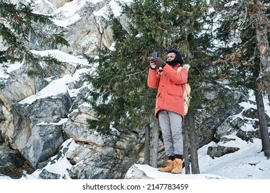 Long Shot Of Young Black Man Hiking In Mountains On Winter Day Standing On Rock Taking Pictures On Smartphone Camera