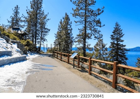 a long smooth concrete walking path with a wooden fence near vast deep blue lake water with lush green trees and plants with snow and blue sky at Lake Tahoe Nevada State Park in Incline Village Nevada