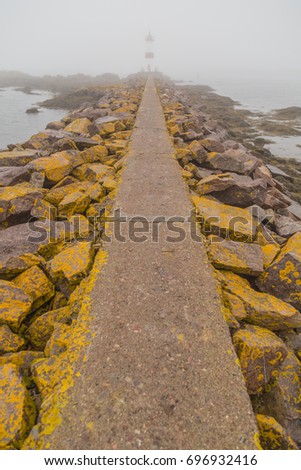 Long stone pathway with large boulders on both sides covered in yellow moss on a foggy day, leading up to a lighthouse on the island of St Pierre and Miquelon.