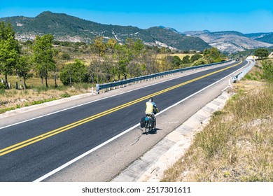 A long, straight highway stretches through a rugged landscape, with a cyclist and vehicles in the distance, symbolizing adventure and freedom on the open road. - Powered by Shutterstock