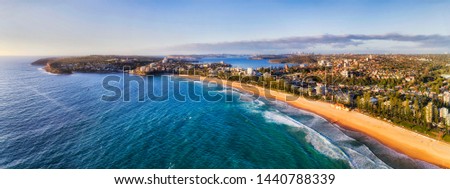 Long strip of clean yellow sandy beach of Manly - part of Sydney famous Northern Beaches seen from mid-air with distant Sydney harbour and Manly waterfront.