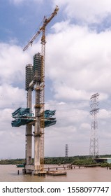 Long Tau River, Vietnam - March 12, 2019: Focus On Tall Suspension Pylon Raising Out Of Brown Water At Phuoc Khanh Bridge Under Construction. Under Blue Cloudscape And Green Belt.