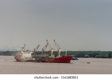 Long Tau River, Vietnam - March 12, 2019: Sea Cargo Vessel, Vissai VCT 05, With Its Own Cranes Anchored In Brown Water. Small River Barge, Along Side, Is Loaded Off Big Ship. Green Belt And Blue Sky.