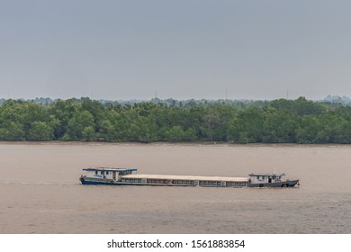 Long Tau River, Vietnam - March 12, 2019: Long Hull-closed River Barge Sails On Brown Water And Lies Deep. Green Belt Separates From Light Blue Sky.