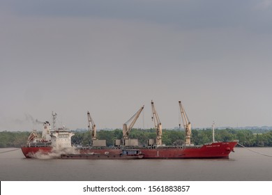 Long Tau River, Vietnam - March 12, 2019: Big Red Sea Cargo Vessel, Vissai VCT 05, With Its Own Cranes Anchored In Brown Water. Small River Barge, Along Side, Is Loaded Off The Big Ship. Green Belt An