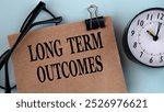 LONG TERM OUTCOMES - words on brown paper on a light background with glasses and a clock. Info concept