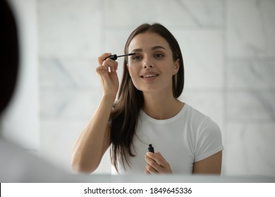Long thick eyelashes. Happy inspired millennial female spending time by mirror making herself up using facial decorative cosmetics holding applicator putting liquid black waterproof mascara on lashes