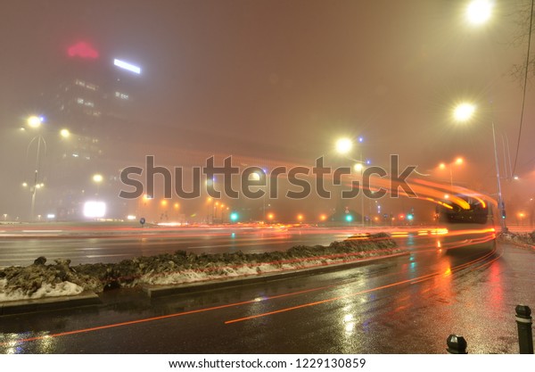 Long traces of the bus tail lights on the misty\
winter evening in the city square with high-rise building and\
street lights.