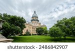 Long view of the Illinois State Capitol Building in Springfield, IL, USA. Cloudy overcast skies overhead. Springtime scene with a lush green lawn and full trees.