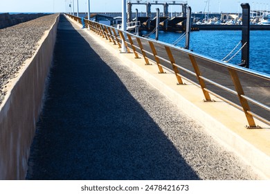 A long walkway with a railing and a dock in the background. The walkway is empty and the dock is full of boats. Concrete walkway stretching into the distance - Powered by Shutterstock