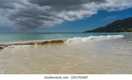 A long wave rolls onto the shore, twisting. Turquoise water mixes with the sand of the beach. Picturesque clouds in the blue sky. A hill in the distance. Seychelles. Mahe.Beau Vallon - Shutterstock ID 2251701487