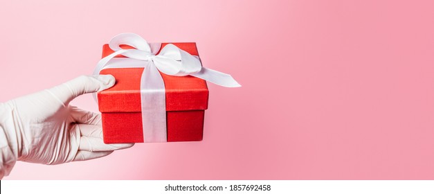 Long Web Banner Side View Closeup Of Human Hand In White Surgical Gloves Holding A Red Gift Box Isolated On Pink Background. Studio Shot