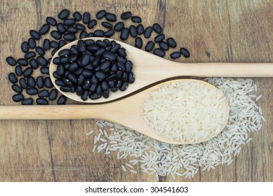 Long White Grain Rice And Black Beans In Wooden Spoons Over A Wooden Background