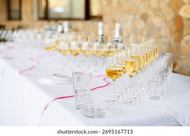 A long white table is lined with numerous champagne and water glasses, pink ribbon accents, and blurred bottles in the background, conveying celebration, hospitality, and refined party atmosphere.