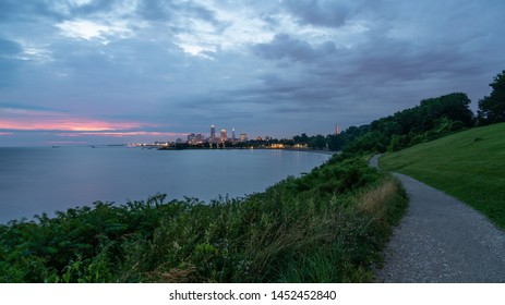 Long Winding Path Along Lake Erie At Edgewater Park With Downtown Cleveland's Skyline In The Distant Background With Overhead Clouds And A Little Sunrise Color.