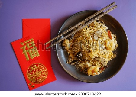 Longevity noodles or stir fried noodle in dark plate with chopsticks on purple background with blurred red envelope with blessing word means ‘good fortune’ Chinese Lunar New Year lucky food (top view)
