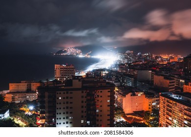 A long-exposure night urban landscape with a coastal Ipanema and Leblon districts of Rio de Janeiro, Brazil, a motion-blurred sky, highly illuminated beach glowing with neon lights, residential houses