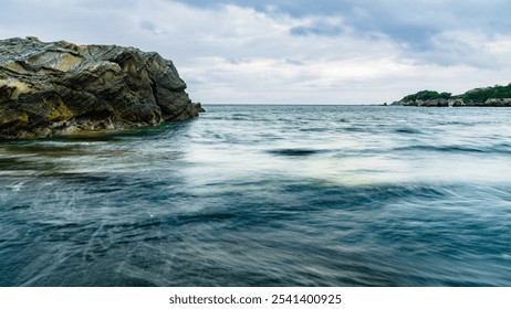 Long-exposure photo capturing rippling waves against the dark sandy shore at Soleil Hill Beach. On the left, a rock formation composed of alternating layers of pyroclastic sandstone and mudstone. - Powered by Shutterstock