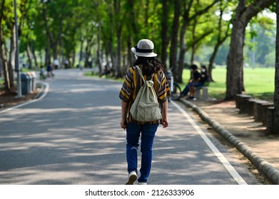 A Long-haired Asian Woman Wearing A Medical Mask Walks On A Walkway In A Shaded Park With Trees During The Epidemic Of COVID-19 Or The Spread Of PM 2.5 Dust. Woman Wear Hats, Jeans, Walking Exercise 