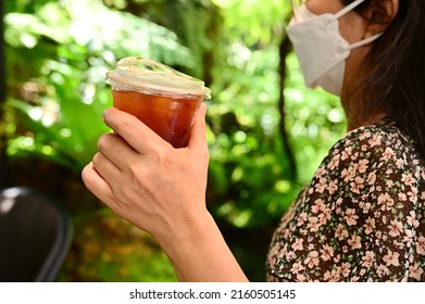 A Long-haired Asian Woman Wearing A White Medical Mask Holds A Cup Of Iced Black Coffee Packaged In A Plastic Cup With A Lift-up Lid.(take Home) With Her Right Hand As She Walks Through The Garden.
