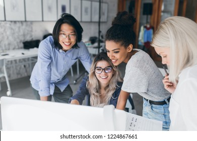 Long-haired Blonde Secretary In Glasses Laughing While Looking At Computer With Asian Programmer And African Girl. Indoor Portrait Of Chinese Office Worker And His Colleagues With Laptop On Foreground