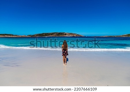 a long-haired girl in a black dress with roses walks along a paradise beach with white sand and turquoise water and orange rocks, cape le grand national park near esperance, western australia