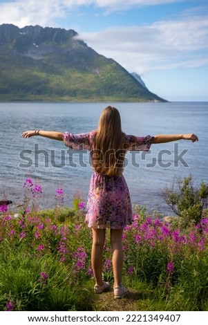 a long-haired girl in a colourful dress enjoys the sunny weather on the island of senja, norway, purple flowers and huge moss-covered mountains in the norwegian cliffs