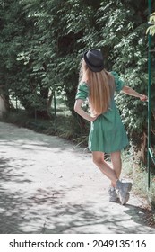 A Long-haired Girl In A Turquoise Dress And A Hat Stands On An Alley In The Park Holding On To A Lattice Bar With Her Hand. Back View