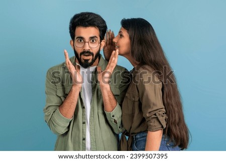 Long-haired indian woman whispering her boyfriend rumors. Shocked millennial eastern guy listening shocking news from his wife, isolated on blue studio background