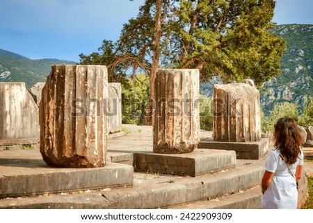 A long-haired woman from behind looking at stones and doric columns of  the ancient temple complex of Athena Pronaia in Delphi. Archaeological site, UNESCO World Heritage Site, Delphi, Greece.