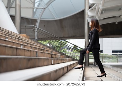 A long-haired woman wearing a mask, suit jacket, trousers and black shoes is walking up the stairs.
