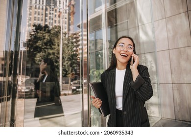 Long-haired Young Asian Brunette Has Cheerful Conversation On Phone Going To Workplace In Office. Active Lifestyle Of Business Woman. Black Formal Suit With Stripes On Her.