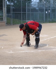 Longmont, CO USA - June 9, 2021: An Umpire In A Red And Black Uniform Cleans Off Home Plate With A Small Brush                              