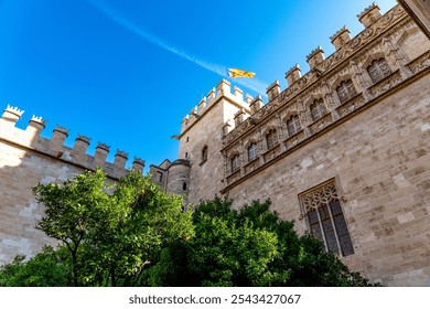 Lonja de la Seda in Valencia, gothic building with ornate windows, spires, flag, blue sky, tree crown, and finely crafted stone reliefs. - Powered by Shutterstock