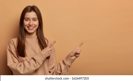 Look, Advertise Here! Dark Haired Pretty Woman With Appealing Smile Gives Positive Attitude Towards Good Deal, Expresses Her Recommendation While Pointing On Right Side, Wears Brown Sweater.