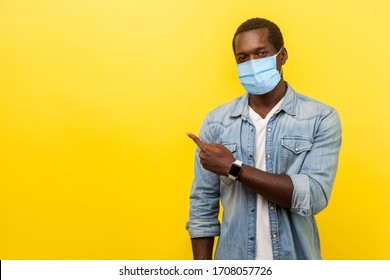 Look, advertise here! Portrait of positive man with surgical medical mask pointing left side and smiling at camera, showing empty space for advertise. indoor studio shot isolated on yellow background