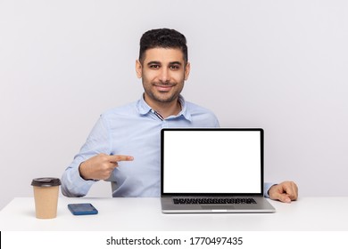 Look At Advertisement! Happy Elegant Man Employee Sitting In Office Workplace, Pointing Laptop Blank Display, Mock Up Empty Place For Website Promotion. Indoor Studio Shot Isolated On White Background