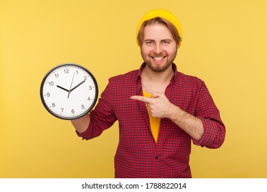 Look At Clock, Don't Rush! Positive Hipster Guy In Checkered Shirt Pointing Big Clock And Looking At Camera With Toothy Smile, Expressing Optimism, Enough Time, Not Hurry. Studio Shot Isolated