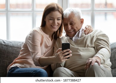 Look, Grandpa. Loving Adult Granddaughter Hugging Smiling Senior Grandfather Taking Selfie With Him On Phone. Young Woman Grown Daughter Embracing Older Man Father Showing Photos Of Grandchild On Cell