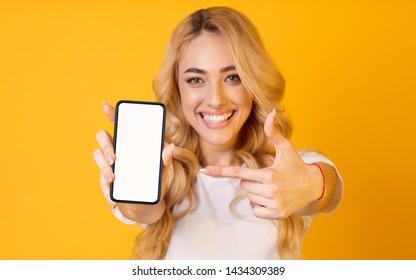 Look Here. Happy Woman Showing Blank Smartphone Screen and Pointing On It, Yellow Background