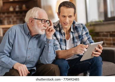 Look here. Pleasant young man pointing at the tablet, showing an interesting article to his elderly father while the man paying attention to him